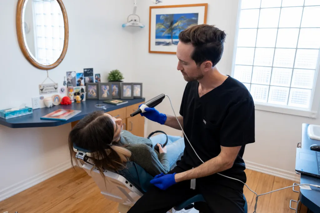 Patient with Dr. Keith during an appointment - Sunrise Dental Arts Dr. Keith, a man with short dark hair in a black scrub, holds a dental tool. A young woman is reclining in the dental chair, holding headphones - Sunrise Dental Arts