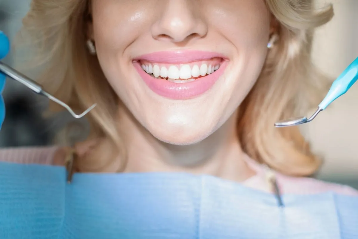 Closeup of beautiful young lady showing her white teeth while having checkup at dental clinic, dentist hands in rubber gloves holding dental tools. Female patient getting teeth whitening treatment