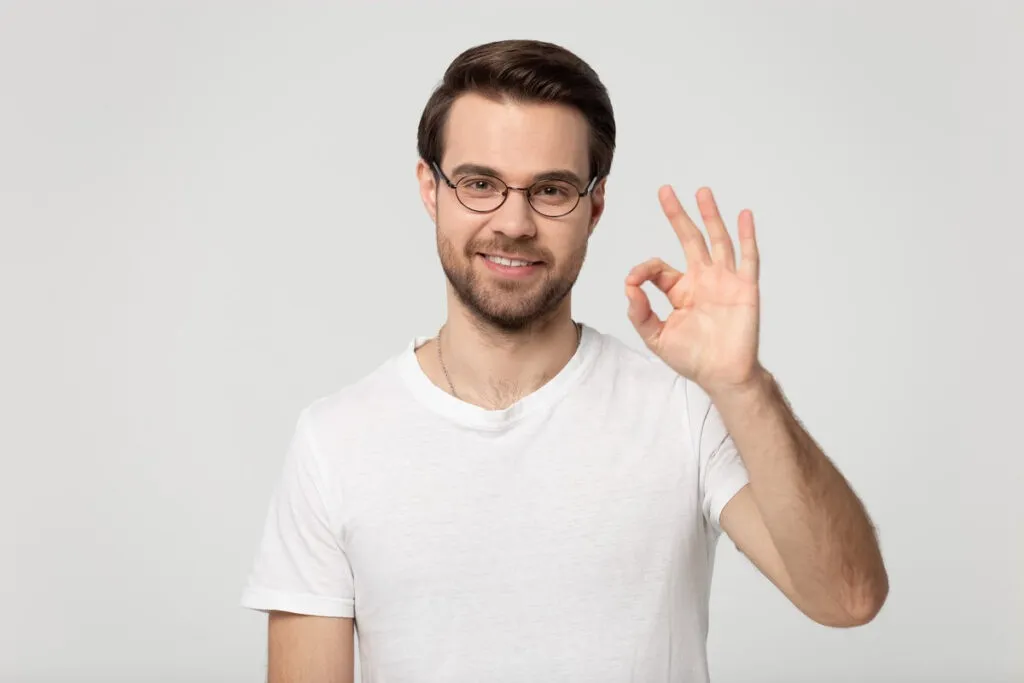 Man pointing at bright smile - Sunrise Dental Arts Care Head shot portrait young happy guy in eyewear showing okay gesture, isolated on grey white studio background. Millennial smiling male client agrees with promotional propose, accepting good offer.