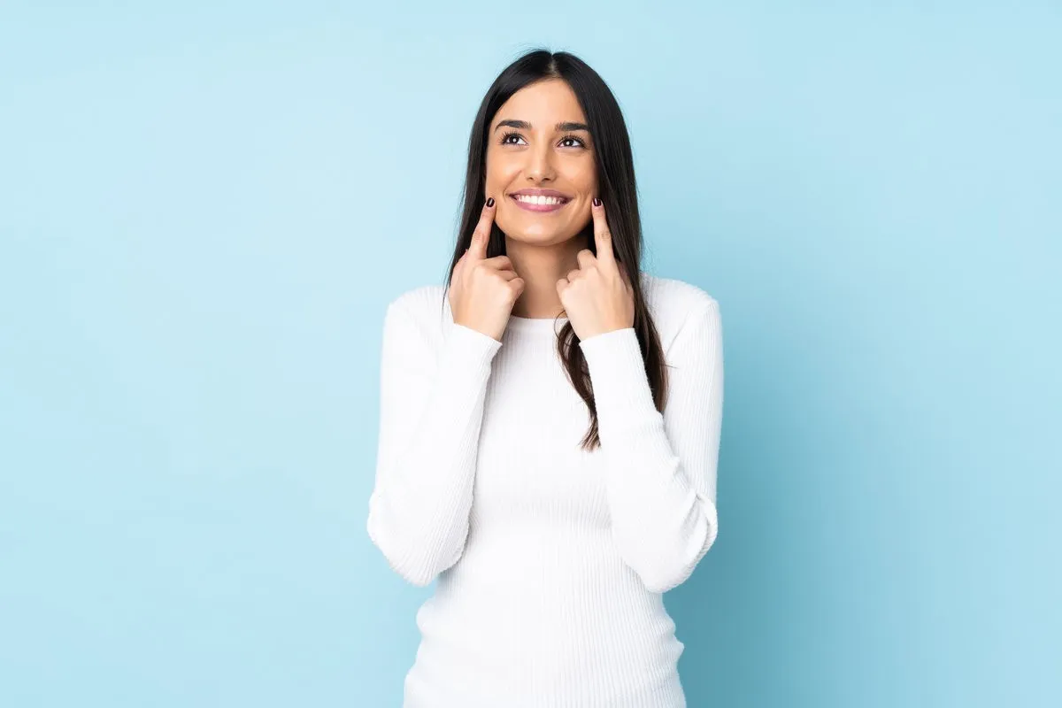 Man pointing at bright smile - Sunrise Dental Arts Care Young caucasian woman isolated on blue background smiling with a happy and pleasant expression