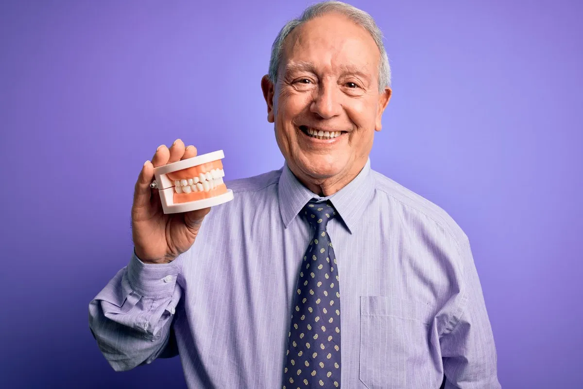 Grey haired senior man holding orthodontic prosthesis denture over purple background with a happy face standing and smiling with a confident smile showing teeth