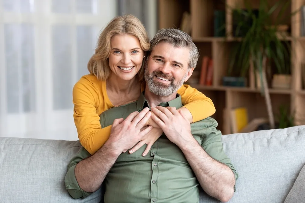 Happy mature couple smiling together - Mission Hills Dentist A joyful middle-aged couple sitting on a sofa, embracing and smiling confidently, showing their bright smiles - Mission Hills Dentist.