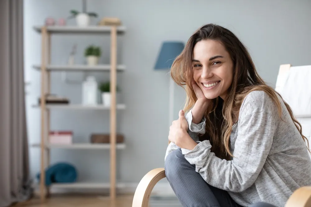 Relaxed woman smiling at home - North Park Dentist A young woman sitting comfortably at home, smiling with confidence and showing her bright teeth - North Park Dentist.