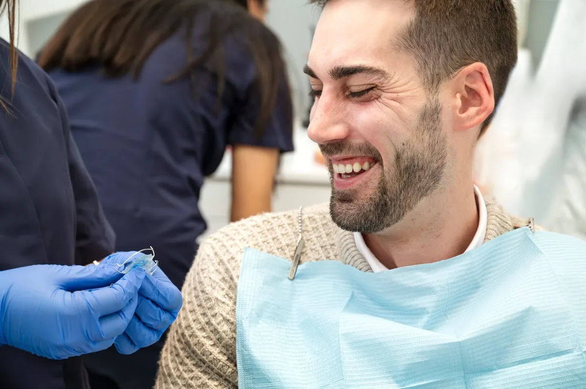 Man pointing at bright smile - Sunrise Dental Arts Care Smiling man in his 30s at a dental checkup with orthodontic retainer in clinic