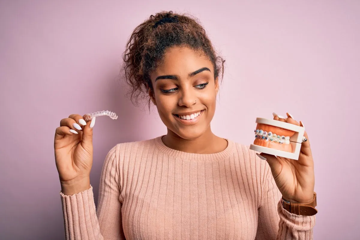 Man pointing at bright smile - Sunrise Dental Arts Care Young african american woman smiling happy holding professional orthodontic denture with metal braces and removable invisible aligner. Comparation of two dental straighten treatments