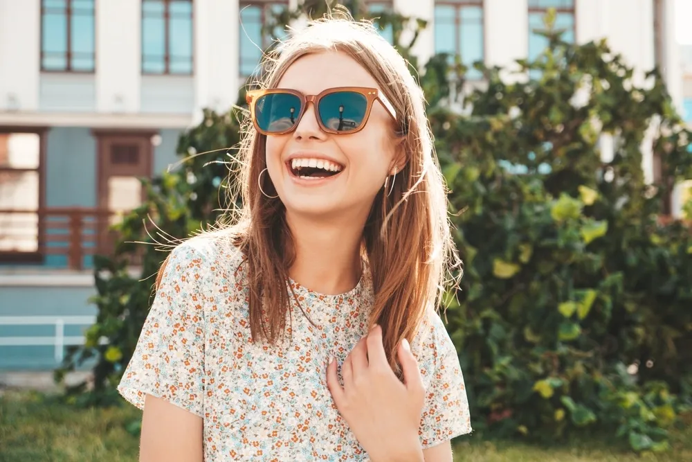 Woman in sunglasses with bright smile - Dentist in Hillcrest A happy young woman wearing sunglasses and a floral top, smiling confidently in the sunshine - Dentist in Hillcrest.