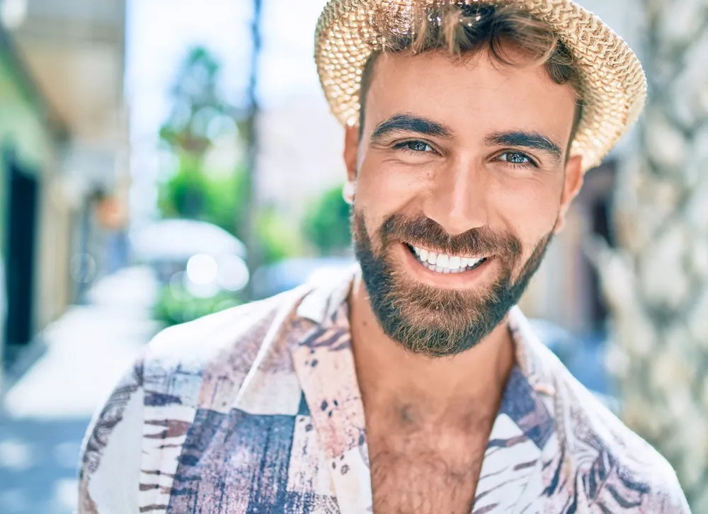 Smiling man in summer hat - Dentist in Hillcrest A cheerful young man with a beard and straw hat smiling brightly outdoors, showing healthy teeth - Dentist in Hillcrest.