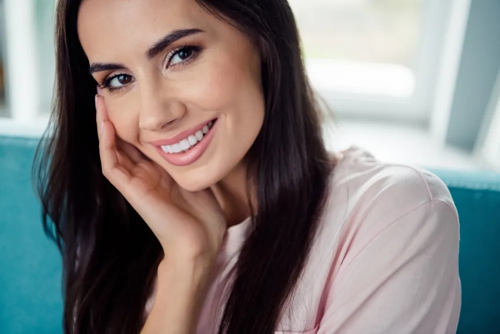 Person sitting on a sofa smiling and touching their cheek after receiving veneers